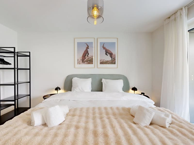 Modern bedroom with white walls, a king-sized bed, two nightstands, framed prints above the bed, a floor lamp, a white curtain, and a black shelf. The bed is neatly made with white bedding and a fluffy beige blanket. The nightstands are accompanied by sma