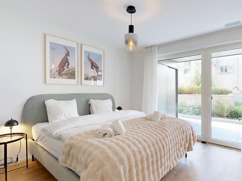 modern bedroom with gray headboard, two single beds with white linens, two white pillows, gray blanket on bed, two lamps, framed pictures of animals, ceiling light, white walls, floor to ceiling windows, balcony with white curtains, terrace with greenery