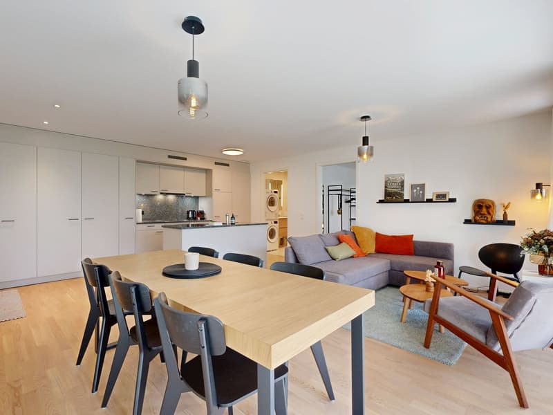 Dining area with a wooden table, open to a kitchen with white cabinets and granite countertops. Living room with a grey sofa, decorative cushions, and a carpet. Modern lamps, an armchair, and a black chair are present. Two side tables, flower vase, and a