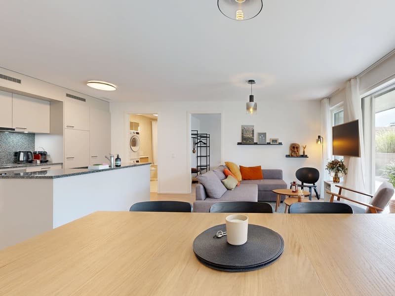 Modern open-plan living room, grey sofa, large dining table, kitchen island with breakfast bar, built-in appliances, glass windows, sliding glass door leading to balcony, white cabinetry, pendant and wall lighting, flat screen TV, plants and flowers on ba