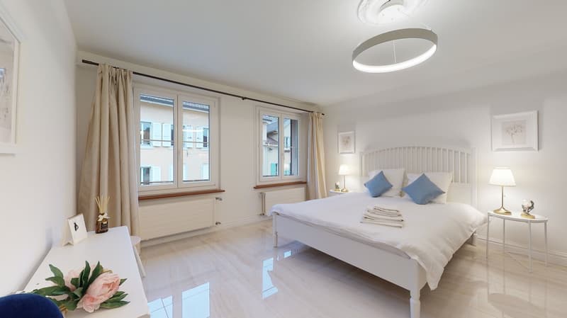 White bed with blue and white pillows, night stands with lamps, flowers on the table, round ceiling lamp, windows with view