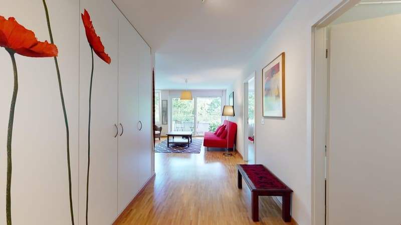 Wide corridor with wooden floor, red sofa in living area, paintings on wall, wooden bench at corridor end, two sets of double doors