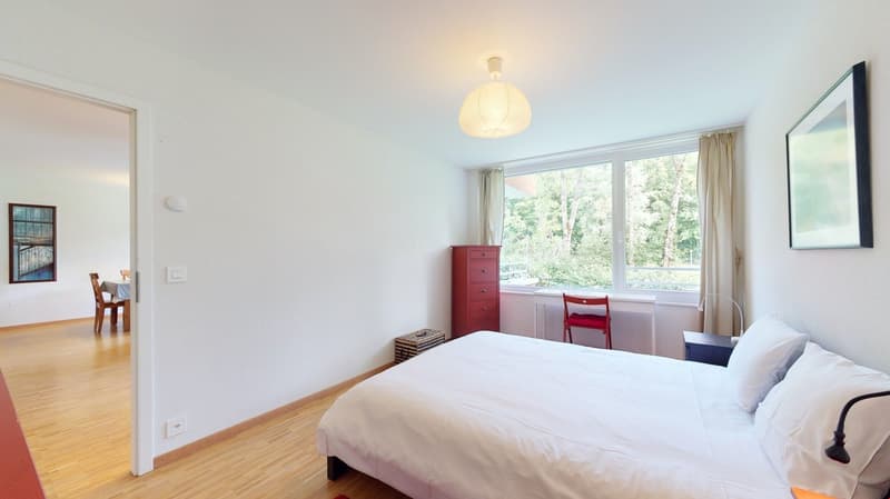 bedroom with wooden floor, white walls, red cabinet, large window, double bed with white sheets, nightstand with bedside lamp, red chair by window, painting