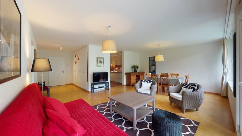 A living room with a red couch, a center table, a TV, lamps, dining table, chairs, a frame, wooden floor, wooden cabinets, kitchen, a flower vase.