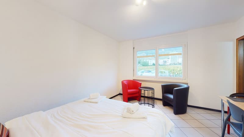 Spacious bedroom with large windows, white walls, tiled floor, red armchair, black armchair, and side tables.