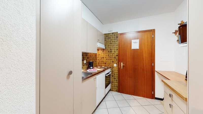 Small kitchenette with white appliances, wooden cabinets, and a tiled backsplash. The room has a tile floor and a wooden door leading to another area.