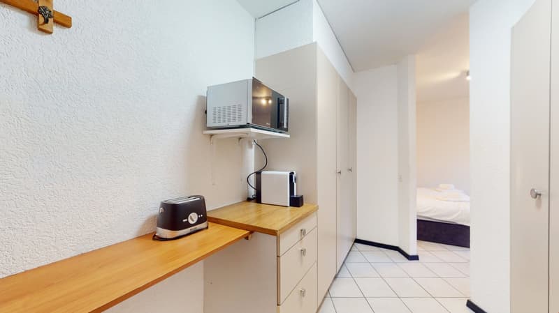 Small kitchenette area with a microwave, coffee maker, and washing machine. Wooden countertop and cabinets. Tiled floor and white walls.
