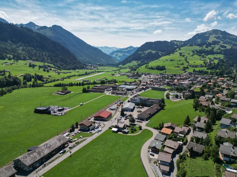 Aerial view of a town with houses, commercial buildings, lush green fields, and mountains in the background. Several cars parked and roads visible.
