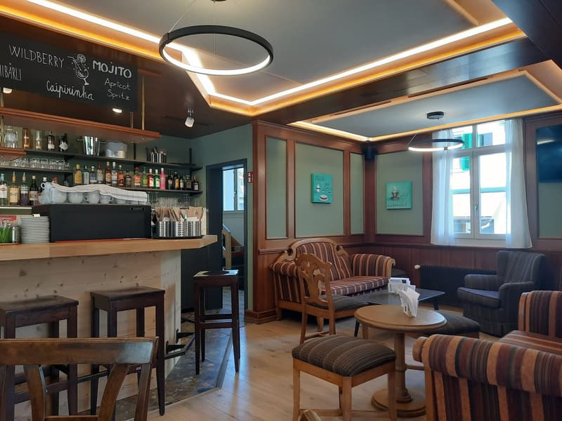 Interior of a pub with a bar counter, stools, chairs, round table, a chalkboard menu, shelves with bottles, a ceiling with round lamps, and paintings on the wall