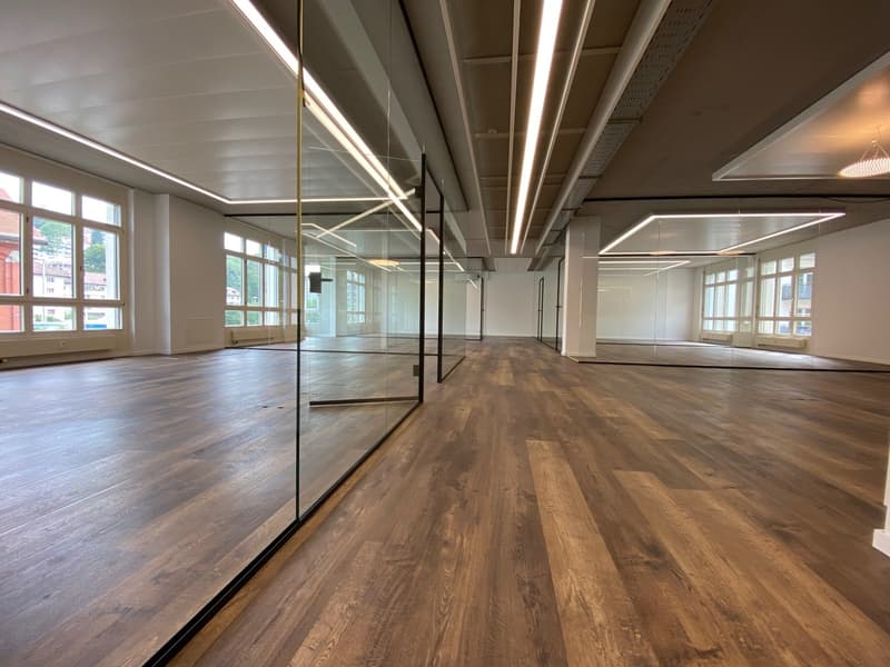 Large open office space with hardwood floors, glass walls, and modern lighting fixtures. The space features tall ceilings, ample natural light from the windows, and a clean, minimalist design.