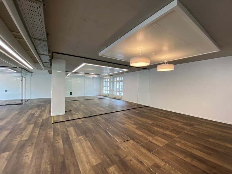 Large room, large windows, mirrors on one wall, wooden floors, two hanging lamps, white walls, ceiling lights.