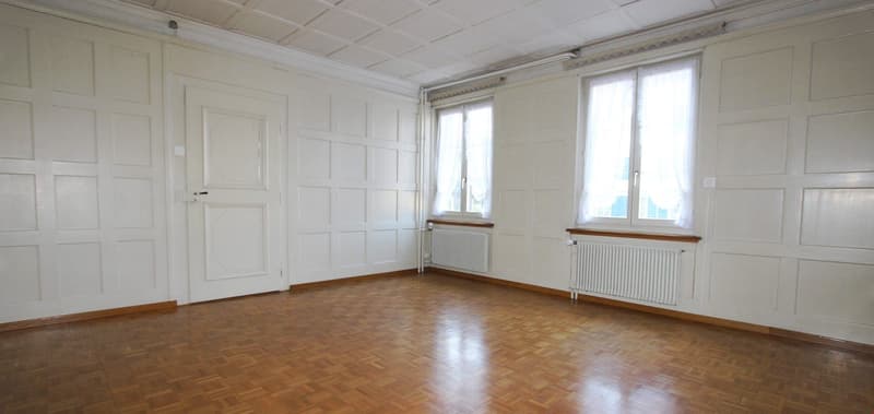 Empty room with wooden floor, white walls, two windows, radiators, and a door.