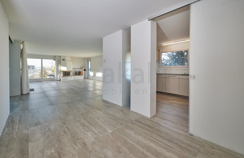 open room with large windows, beige floor, white walls, kitchen with white cupboards and sink