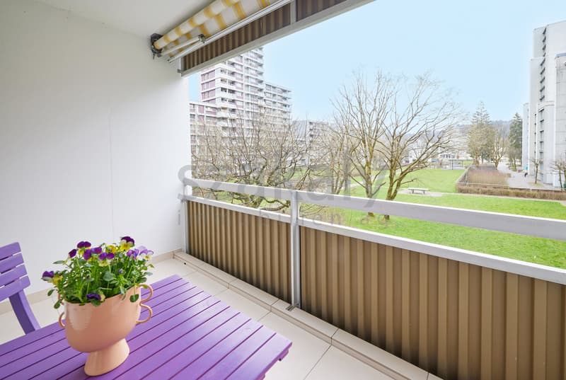 Balcony with table and flowers, white walls, white floor, metal railing, view of a park