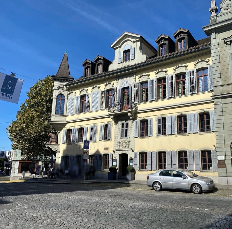 Historic building, yellow facade, blue shutters, multiple balconies, small garden in front, gray cobblestone street