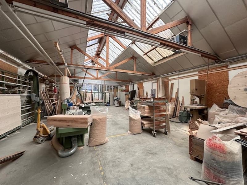 Workshop with high ceilings, wooden trusses, shelves, tables, tools, wooden beams, glass windows, sacks of materials, a trolley, and a pile of plywood.