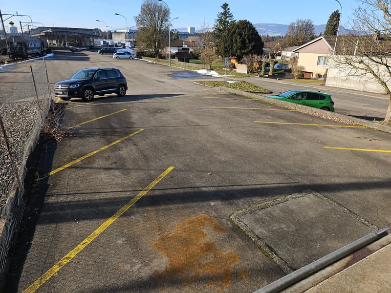 Wide parking lot, clear skies, yellow markings, some greenery, nearby houses, trees, mountains in the distance