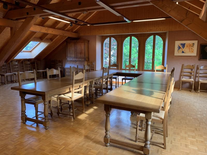 Large open-concept dining room with wooden beams, arched windows overlooking a forested area, wooden dining tables and chairs, and a wooden cabinet or armoire in the corner.