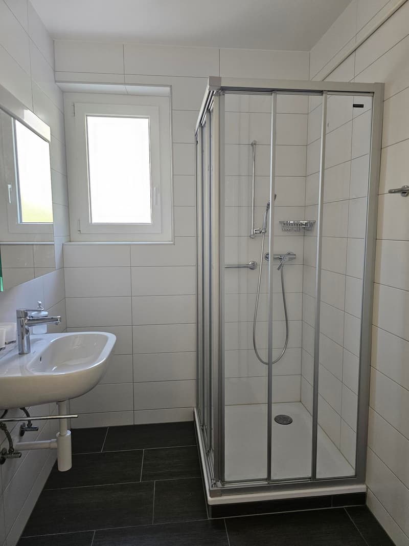 Spacious bathroom with white tiled walls, a glass-enclosed shower stall, a sink, and a window providing natural light.