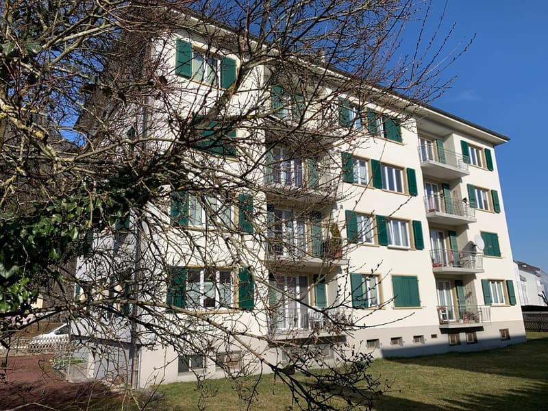 A multi-story apartment building with white exterior walls and green shutters on the windows. The building has several balconies and is surrounded by bare tree branches in the foreground.