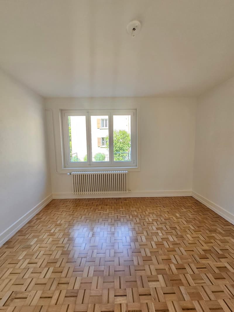 Empty room with white walls, hardwood parquet flooring, and a window overlooking the exterior.