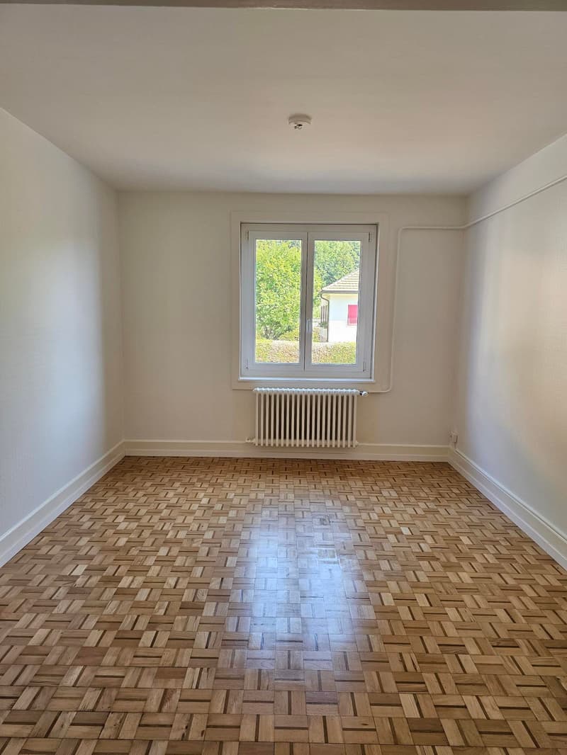 Empty room with white walls, hardwood parquet flooring, and a window overlooking the outdoors.