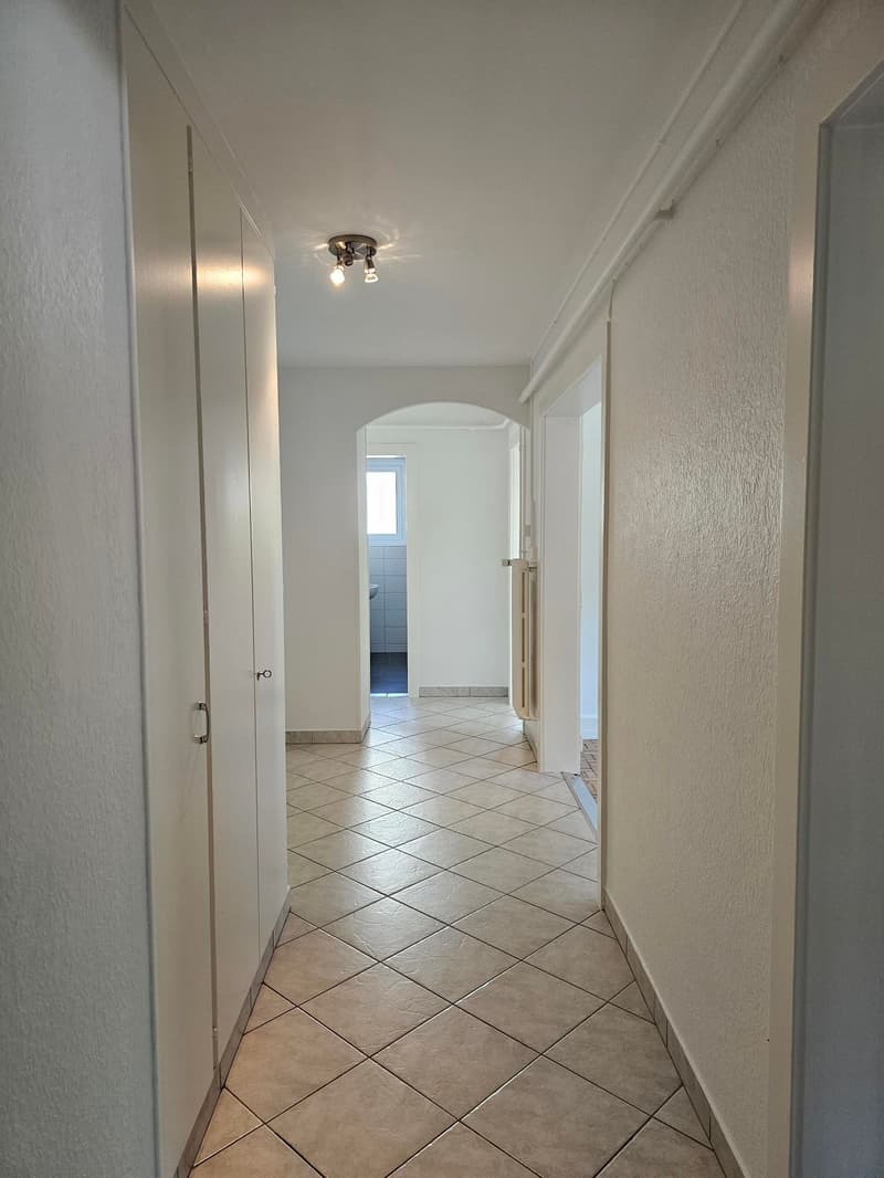 The image shows a hallway with white walls and a tiled floor in a diagonal pattern. There are several doors leading to other rooms, and a light fixture on the ceiling.