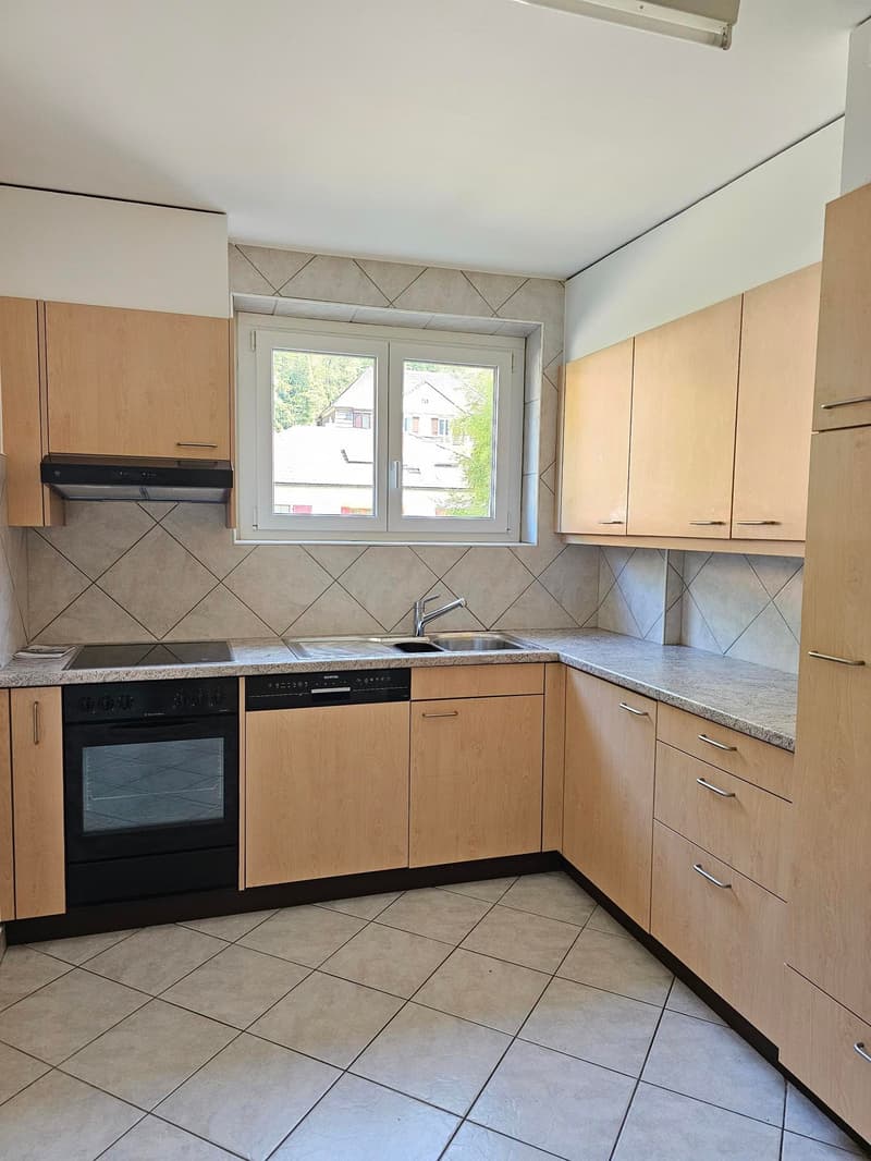 The image shows a modern kitchen with light wood cabinets, gray tile flooring, and a window overlooking the outdoors. The kitchen features a built-in oven, a sink, and countertops.
