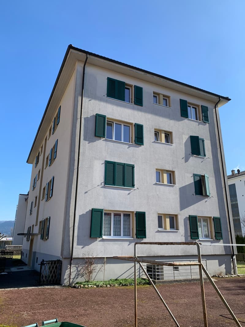 4-story apartment building with a stucco exterior, green shutters, and multiple windows. The building appears to be situated in a residential area with a grassy area in the foreground.
