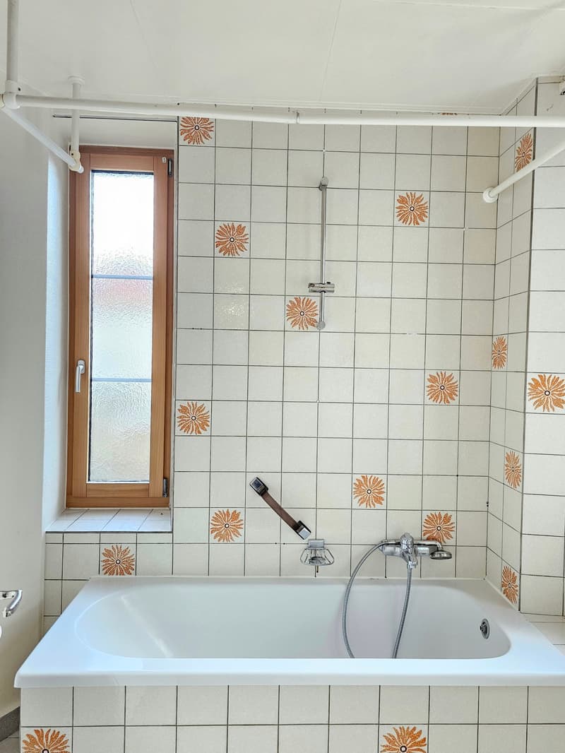 White tiled walls, brown wooden window frame, white bathtub, silver shower head and handle, clear glass window