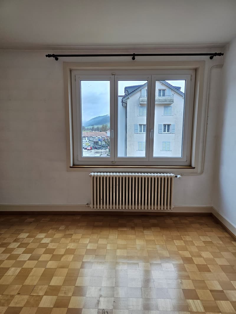 Empty room with wooden parquet flooring, white walls, large window with view to the outside, white radiator