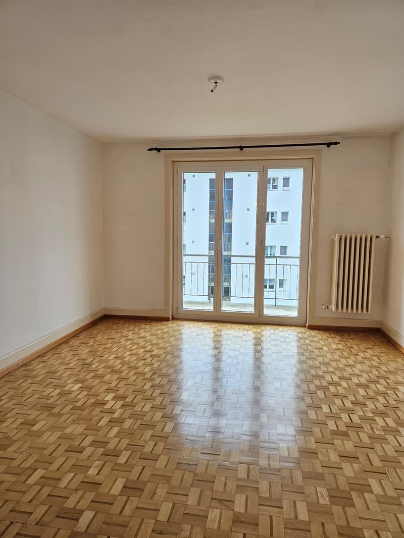 Large empty room, white walls, wooden parquet flooring, sliding glass door leading to balcony with metal railing, white radiator on the wall
