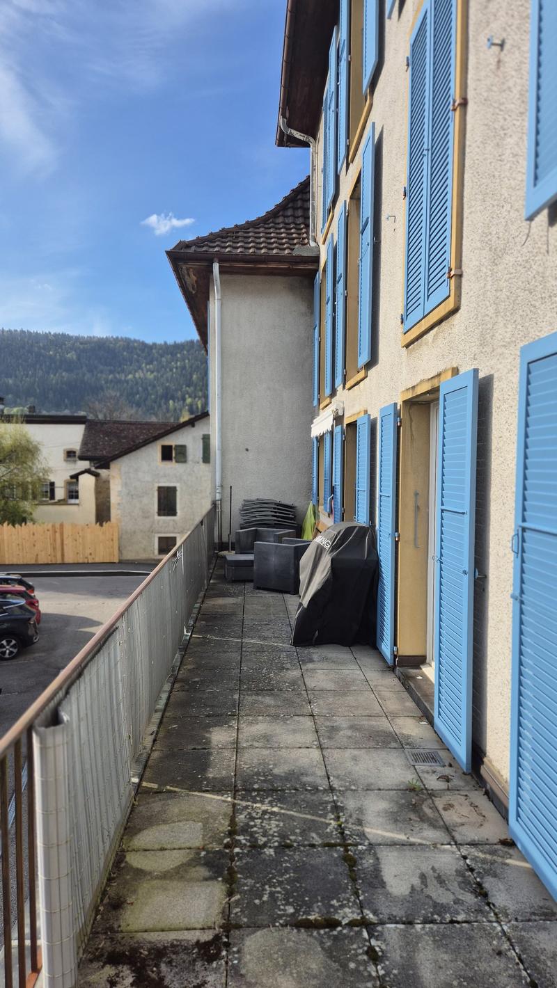 Building with blue shutters, tiled walkway, metal railing, covered outdoor seating, covered grill, cars parked in front