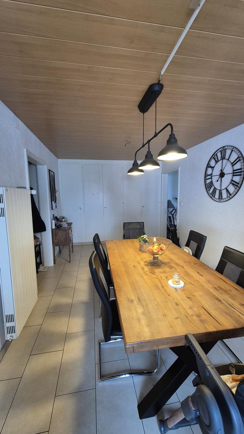 Dining room with wooden table, black chairs, wooden ceiling, tiled floor, white walls, three hanging lamps, clock on wall, wooden cabinet, and a bowl of fruit on the table.
