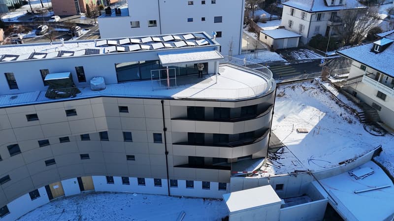 Exterior view of a modern, multi-story residential building with an arched rooftop terrace and a glass-walled entrance. The building has solar panels installed on the rooftop, a white exterior, and is surrounded by a snowy landscape.