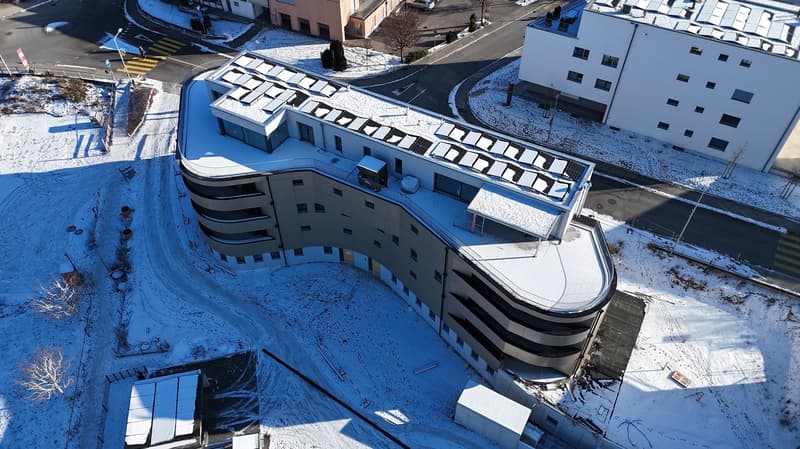 Contemporary multi-story building with balconies, curved design, raised ground floor, solar panels on the roof, snow-covered surroundings