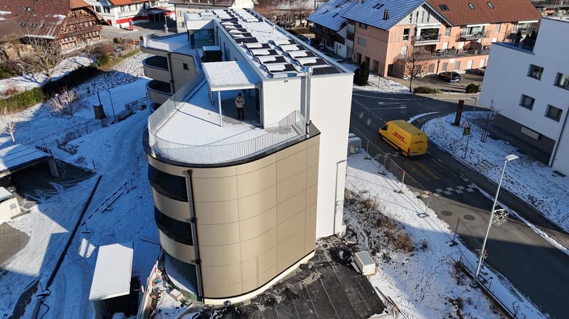 Large building with many windows, curved facade, snow on the ground, road on one side, construction barriers, solar panels on roof