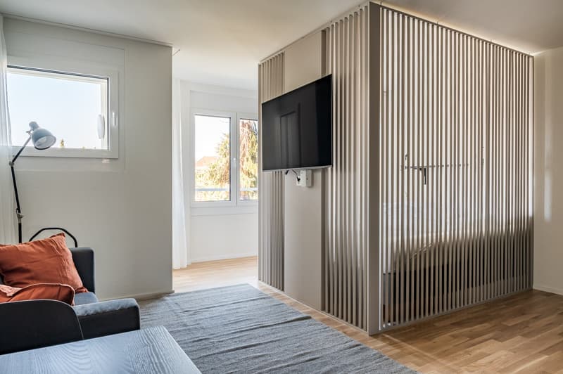 Small room with couch, a table, a mounted television, a window, a lamp, and a curtain.