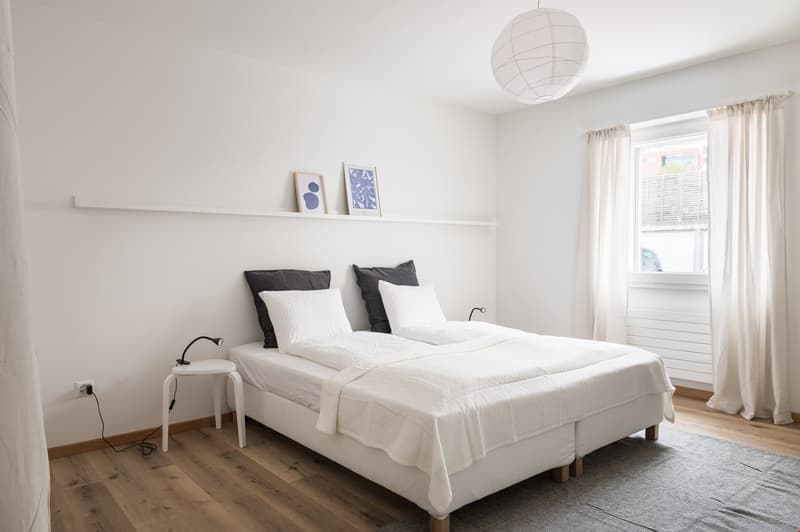 white walls, wooden floor, a double bed, two pillows, white sheets, bedside table, two frames above the bed, round ceiling light