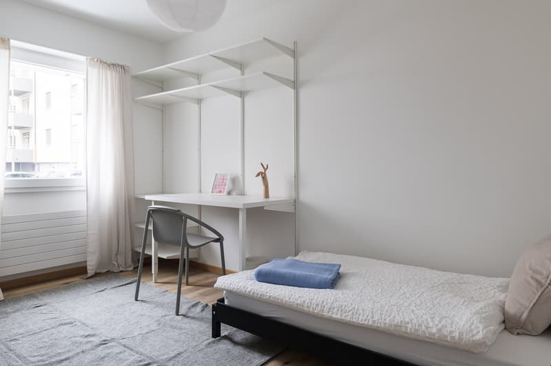 A small bedroom with a desk, chair, and bed. A rug is on the floor. There is a window with white curtains. Above the bed are white shelves with some decorations.