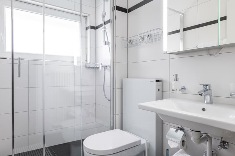 White tiled walls, clear glass shower enclosure, white ceramic sink, modern showerhead, wall-mounted soap dispenser, toilet