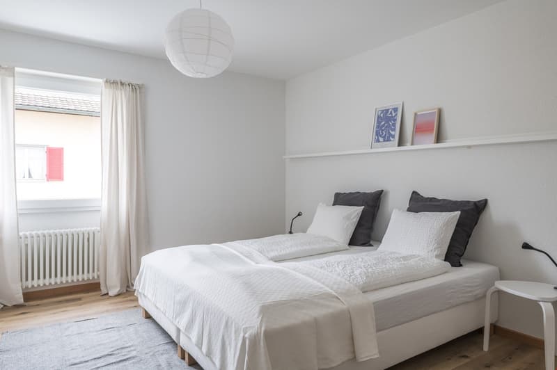 white walls, wooden floor, double bed with white bedding, nightstand, radiator, shelf with picture frames, lamp