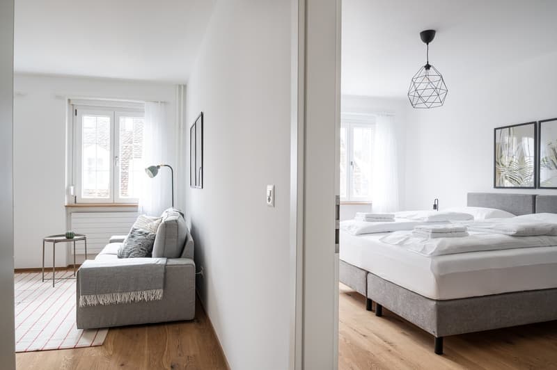 Spacious bedroom with large windows, hardwood floors, gray sofa, and a geometric pendant light. The bedroom leads to an adjacent living area with a balcony or terrace.
