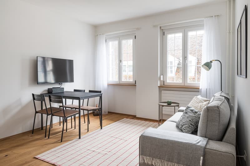 Spacious living room with large windows, hardwood floors, a dining table with chairs, a gray sofa, and a TV mounted on the wall. The room has a minimalist and modern design aesthetic.