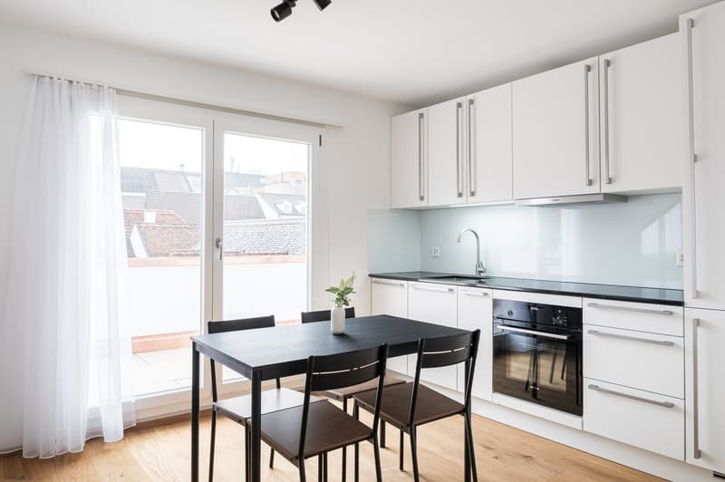 Modern kitchen with white cabinets, black countertop, built-in oven, and dining table with black chairs. Large windows provide natural light and views of the surrounding buildings.