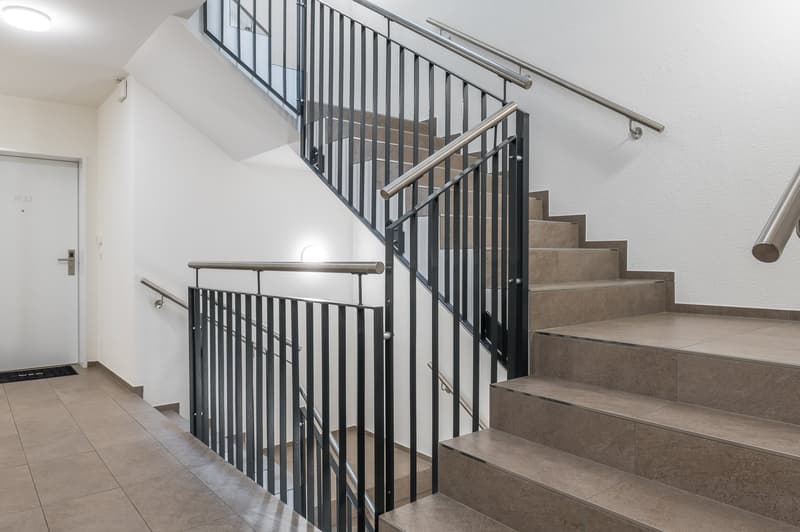 A modern staircase with tiled steps, metal handrails, and a white door to the left. The wall and ceiling are white.