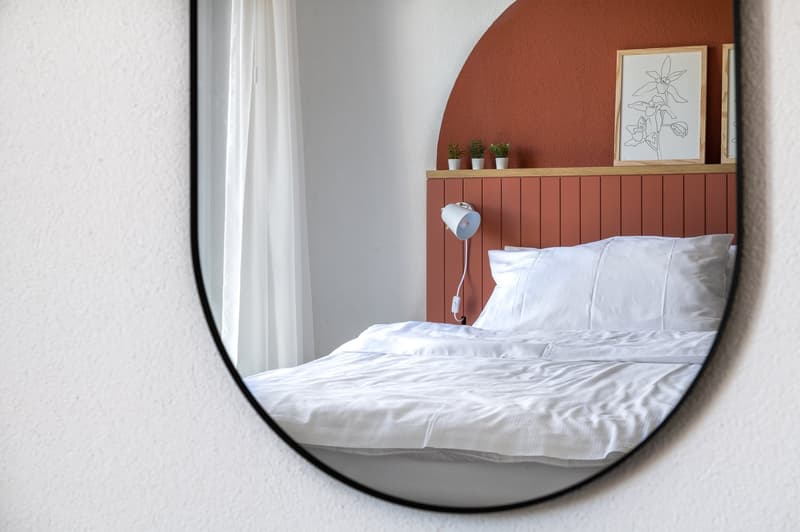 Bedroom with round mirror, white wall, white bedding, red headboard, nightstand lamp, wall decor