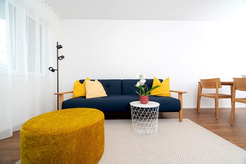 modern living room, blue sofa, yellow pillows, wooden table, potted flower, wooden floor, floor lamp, white curtains