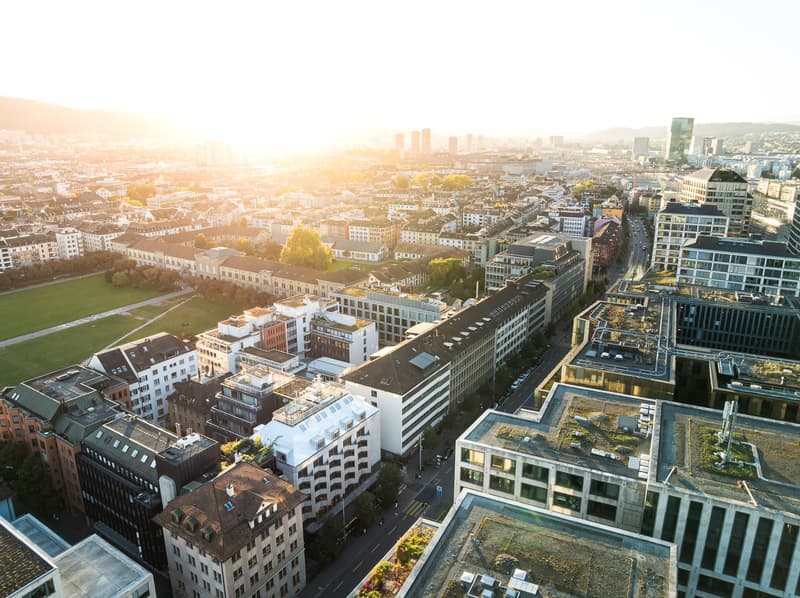 An aerial view of a cityscape with multiple buildings, a wide road, and a large park area. The sun is setting on the horizon, and the buildings are of various sizes and styles. The image features both urban and green spaces.