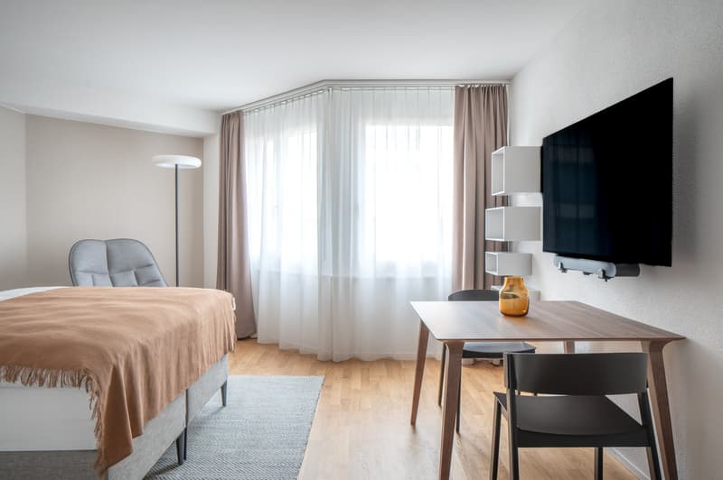 small bedroom with bed, desk, 2 chairs, TV mounted on the wall, rug, floor lamp